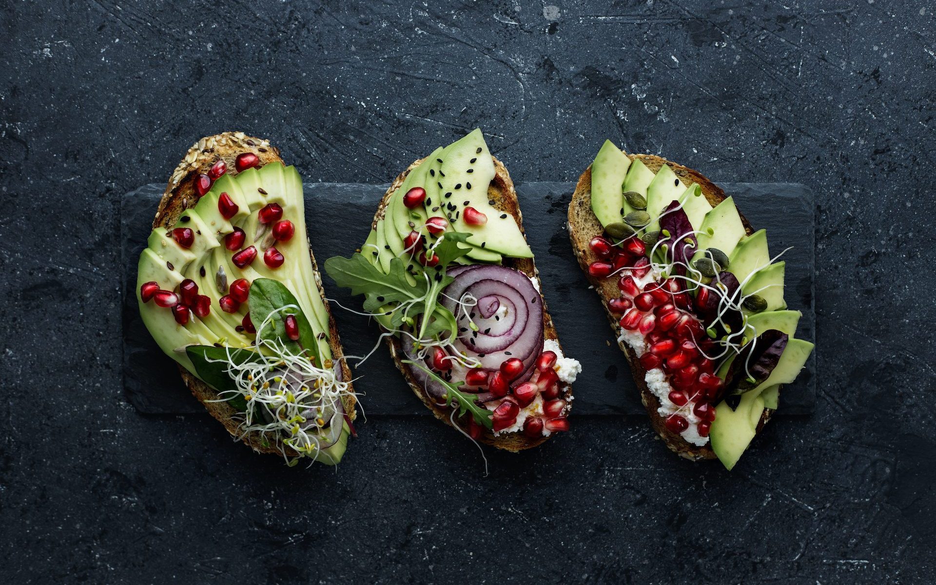 Healthy avocado toasts with avocado slices, pomegranate seeds and sprouts on dark background.